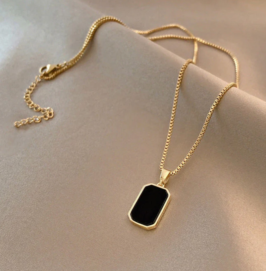 A gold-tone chain necklace with a black rectangular pendant, displayed on a beige fabric surface.